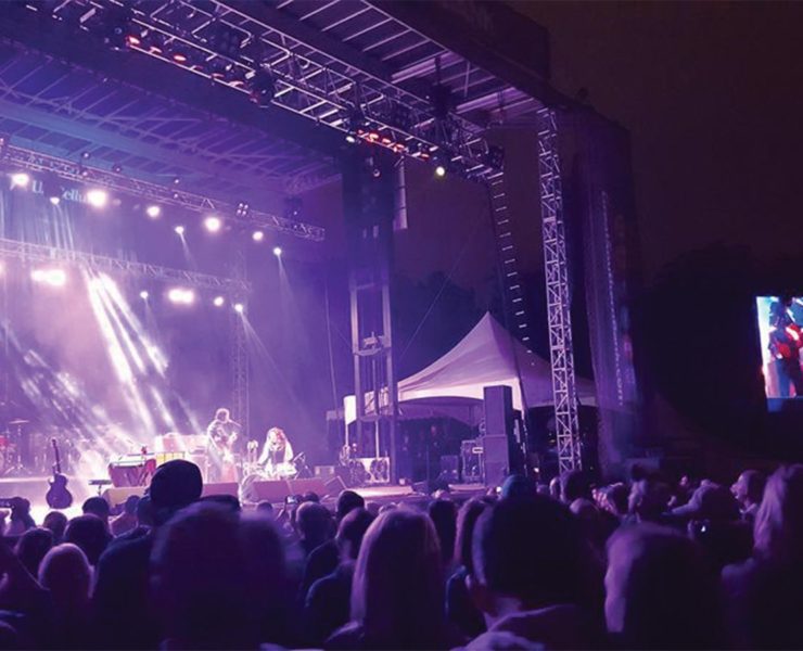 A nighttime concert scene with a band performing on a lit stage. The audience watches, silhouetted in the foreground. Bright spotlights illuminate the performers. A large screen shows the performance to the side.