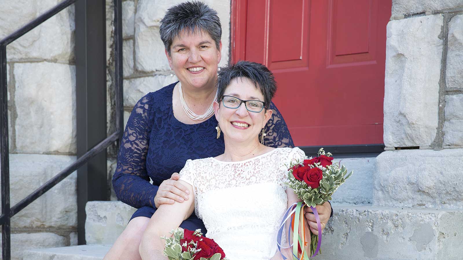 Two women sitting on stone steps in front of a red door. The woman in front wears a white dress and holds red roses, while the woman behind wears a navy blue lace dress. Both are smiling.