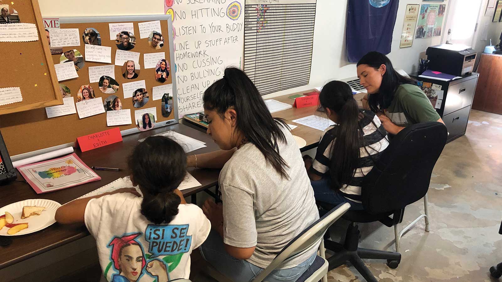 A group of four children work at a long desk with papers, accompanied by an adult who is assisting them. Notes and photos are pinned to a bulletin board on the wall. Snacks are on the table. The room has a casual, educational atmosphere.