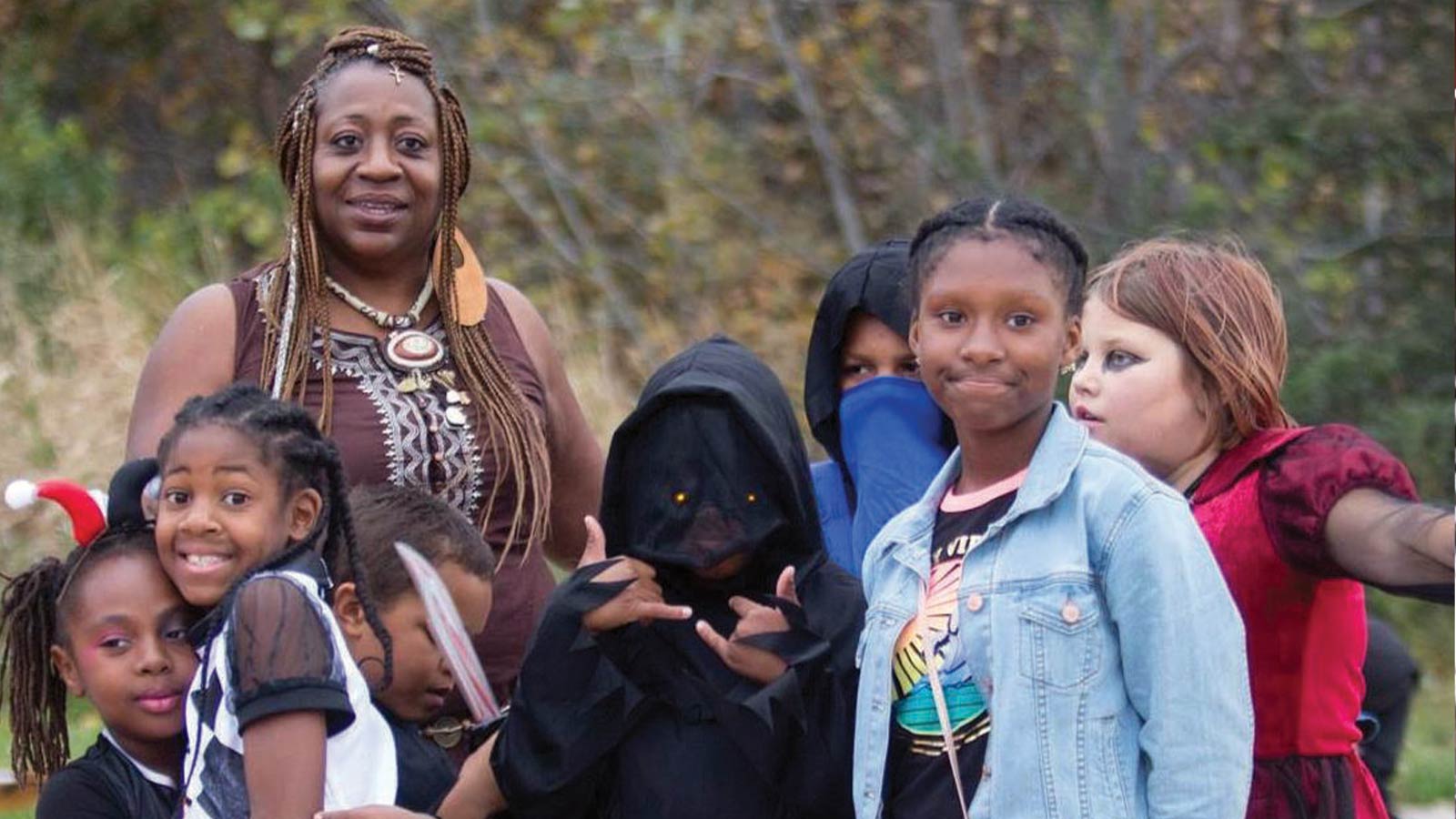 A group of children in Halloween costumes stand outside with an adult. The kids are dressed as various characters, including a hooded figure and a girl in a denim jacket, smiling. Trees and greenery are in the background.