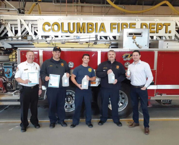 Five men stand in front of a fire truck labeled Columbia Fire Dept. Three are in uniform holding certificates and bottles, while another man in a uniform and a man in a white shirt and jeans smile next to them. They are inside a fire station.