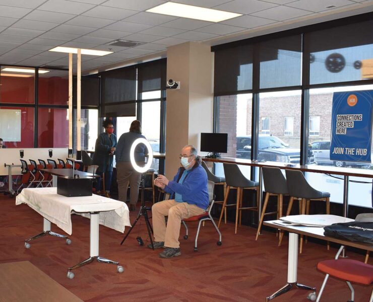 A man wearing a mask is setting up a camera and ring light in a room with large windows. The room has tables, chairs, and a sign on the window that says, Connections Recorded Here. Join the Hub. Another person stands nearby.