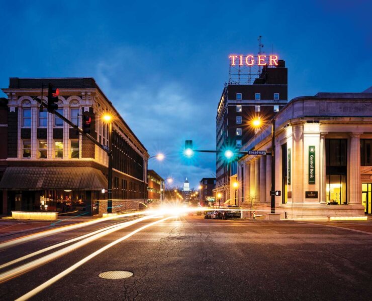 Street scene at dusk with light trails from vehicles. Historic buildings line the road, including one with a large TIGER sign on top. The sky is a deep blue, and streetlights are illuminated, adding a warm glow.