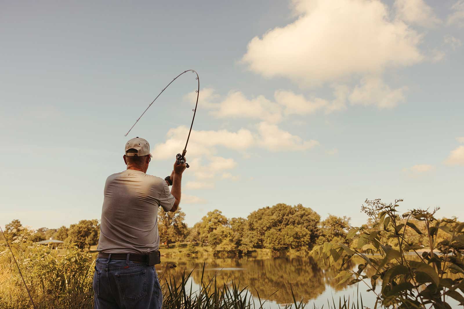 A older man reeling in his fishing rod by the edge of a pond on a sunny day outside