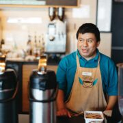 A male barrista wearing a tan colored apron behind the counter of a coffee shop