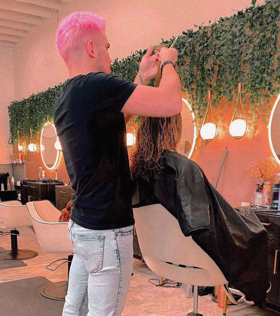 A hair stylist cutting a women's hair at a place called Foxy Boss Salon featuring pink walls, cascading ivy from the ceiling