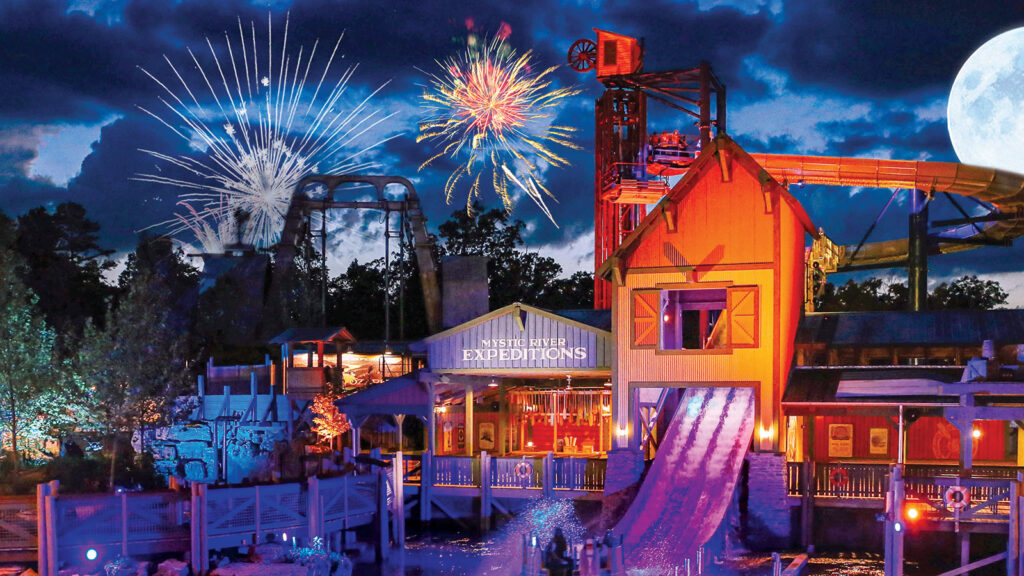 a water themed roller coaster with a pond at night with fireworks lighting up the sky in the background. It is called 