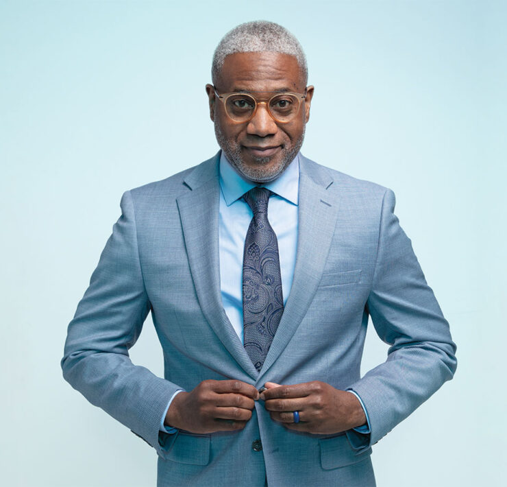 A headshot of a older man named Dr. Lester Woods, Jr. buttoning the front of his blue suit jacket standing in front of a blue background