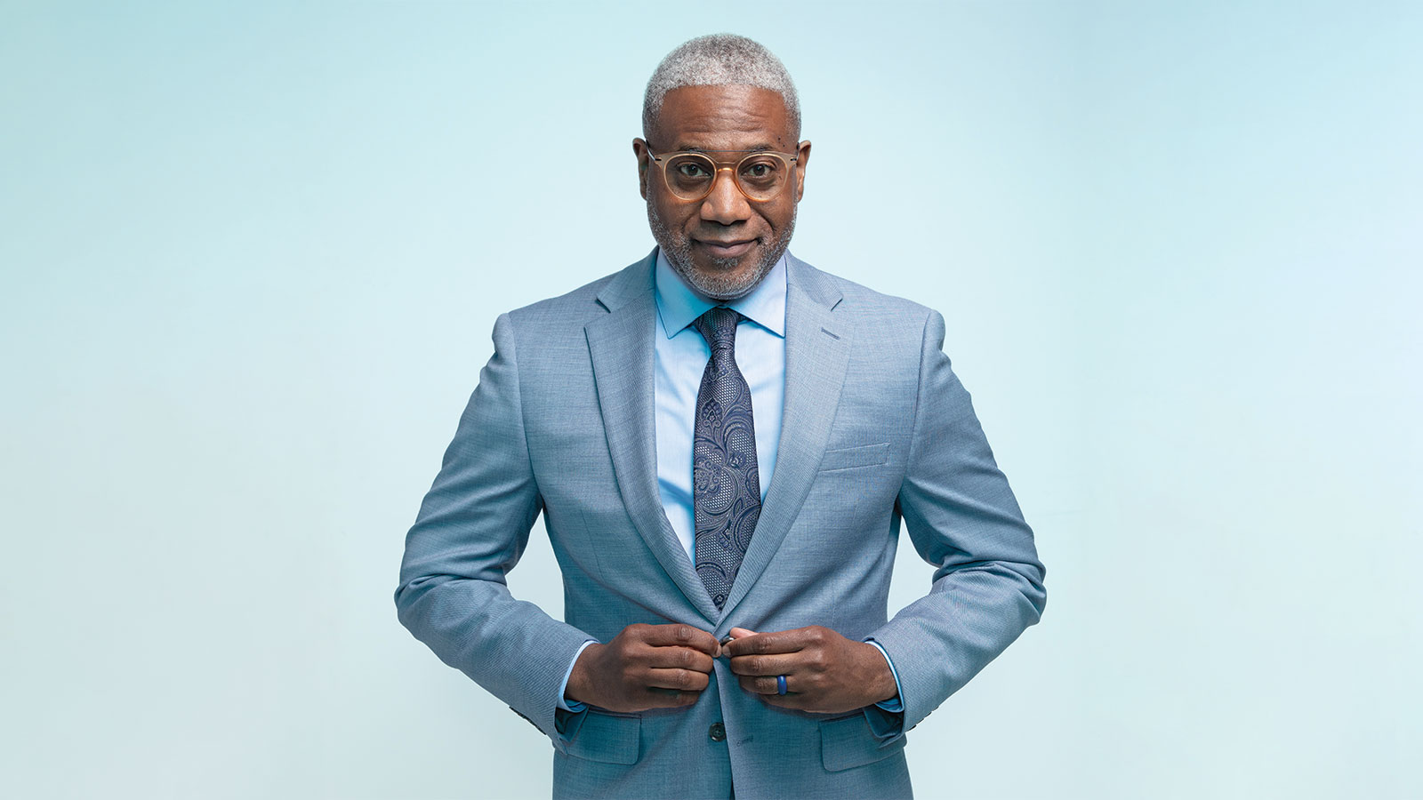 A headshot of a older man named Dr. Lester Woods, Jr. buttoning the front of his blue suit jacket standing in front of a blue background