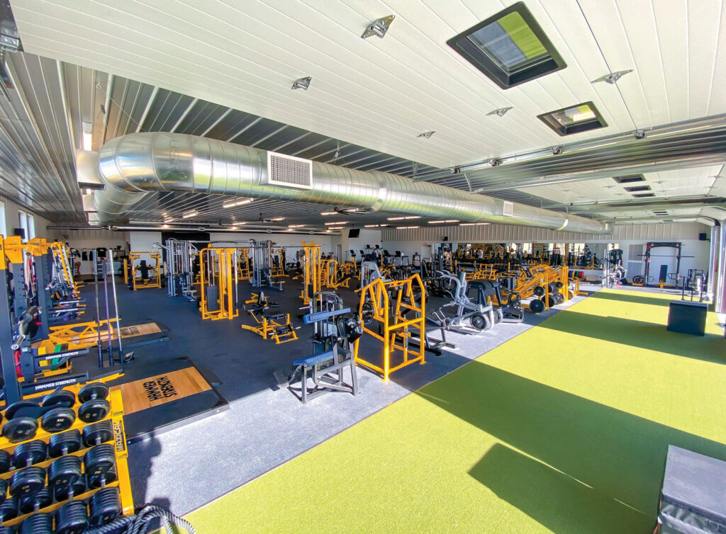 interior of a gym filled with a wide assortment of fitness equipment that is orange and silver colors with part of the ground covered in artificial short turf