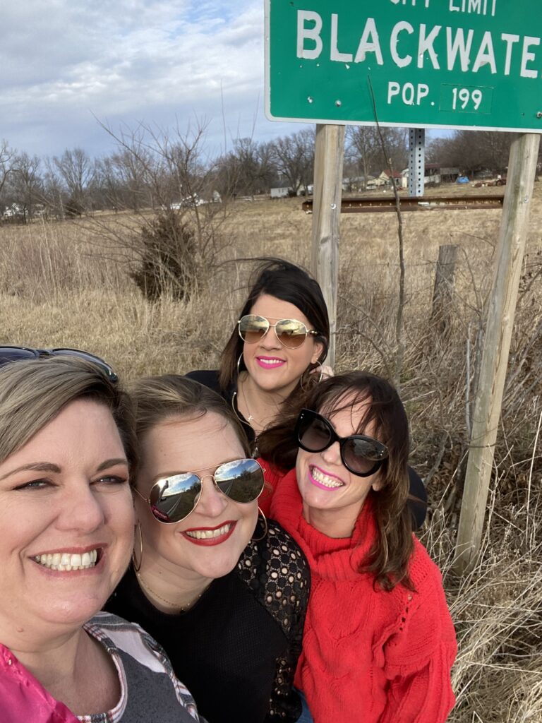 four women pose by a town entrance sign on the side of a dead field