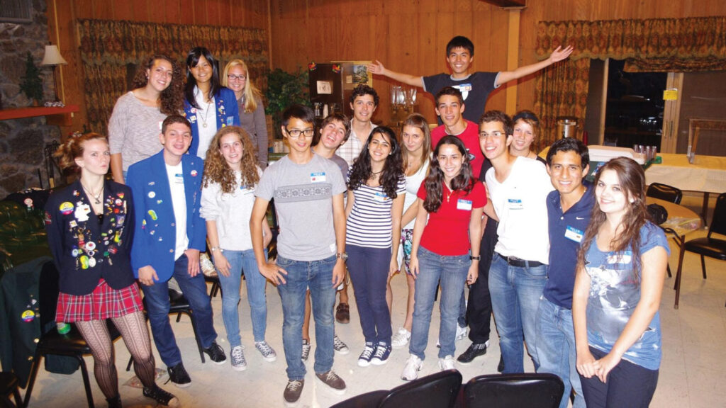 a large group of international students pose in a rustic living room with a stone fireplace and wood paneling on the wall