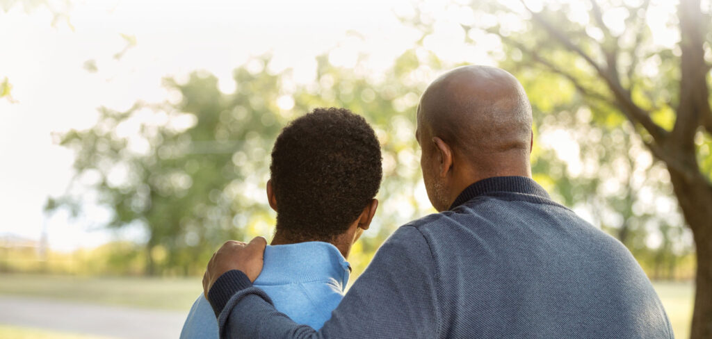 view from the back as a man puts his hand on the shoulder of a teenage boy as they overlook a park