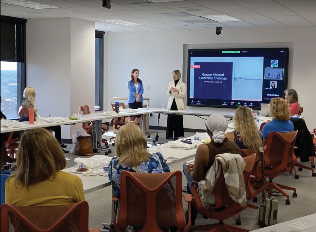 Two presenters stand before a board room table filled with women