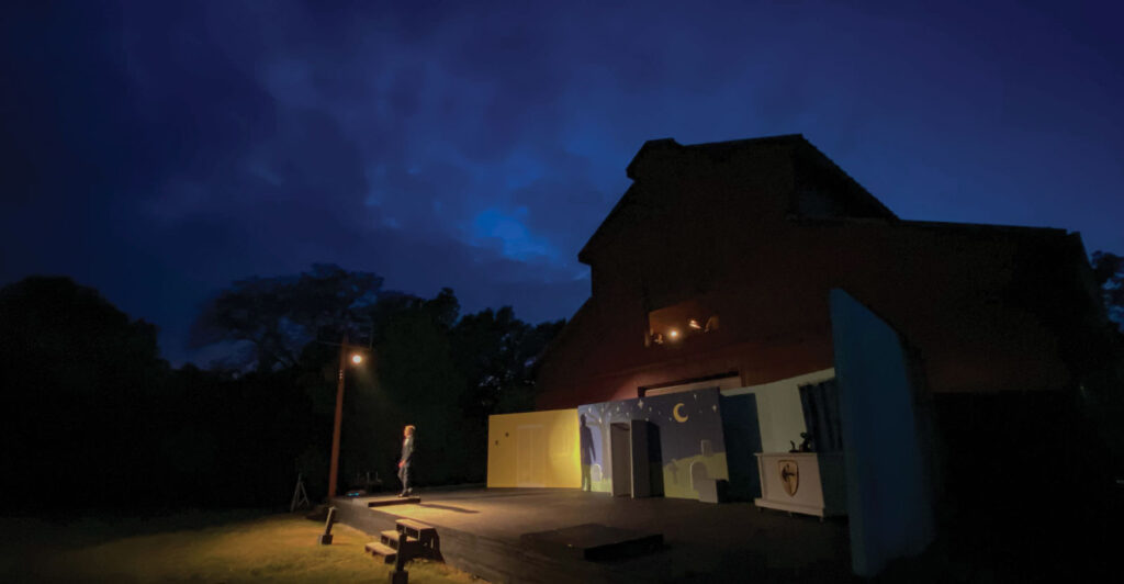 stage in front of a red barn at night with one light focused on one young performer on the stage