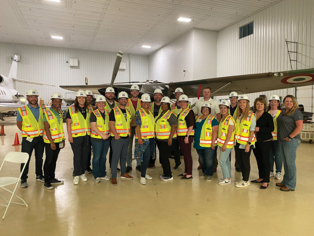 A group of educators pose with students in hard hats and reflection vests inside of a airplane hanger