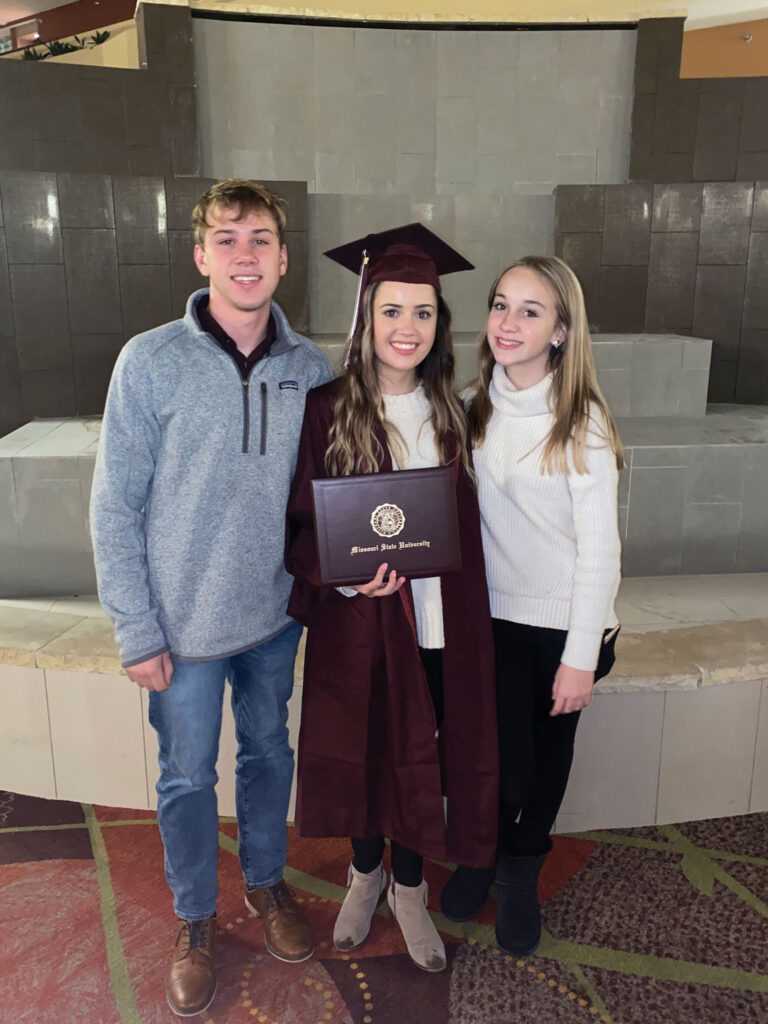 a young women in a graduation cap and gown pose alongside young girl and boy