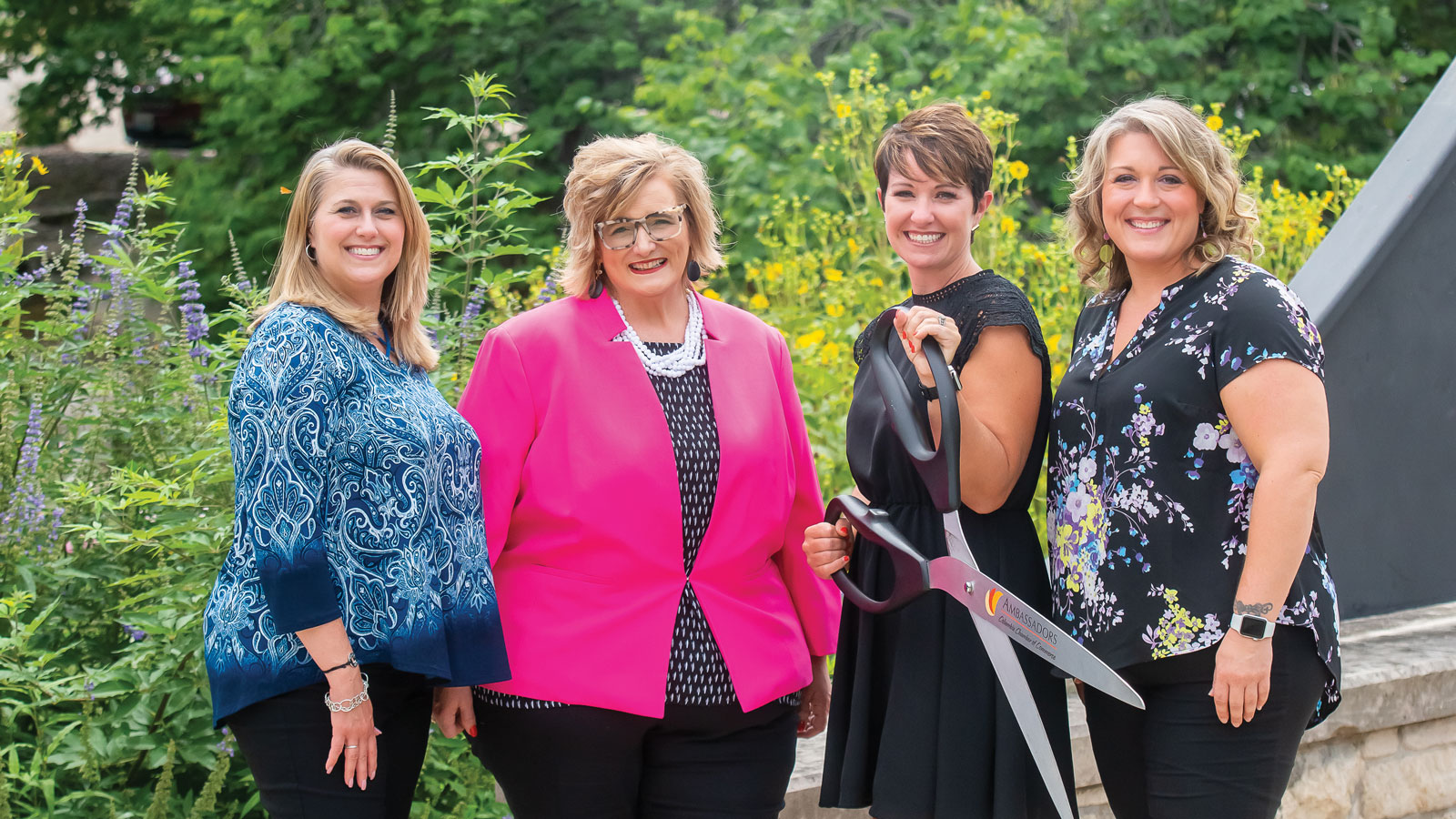 Close up shot of four women apart of Columbia chamber of commerce stand outside near a garden holding a large pair of scissors