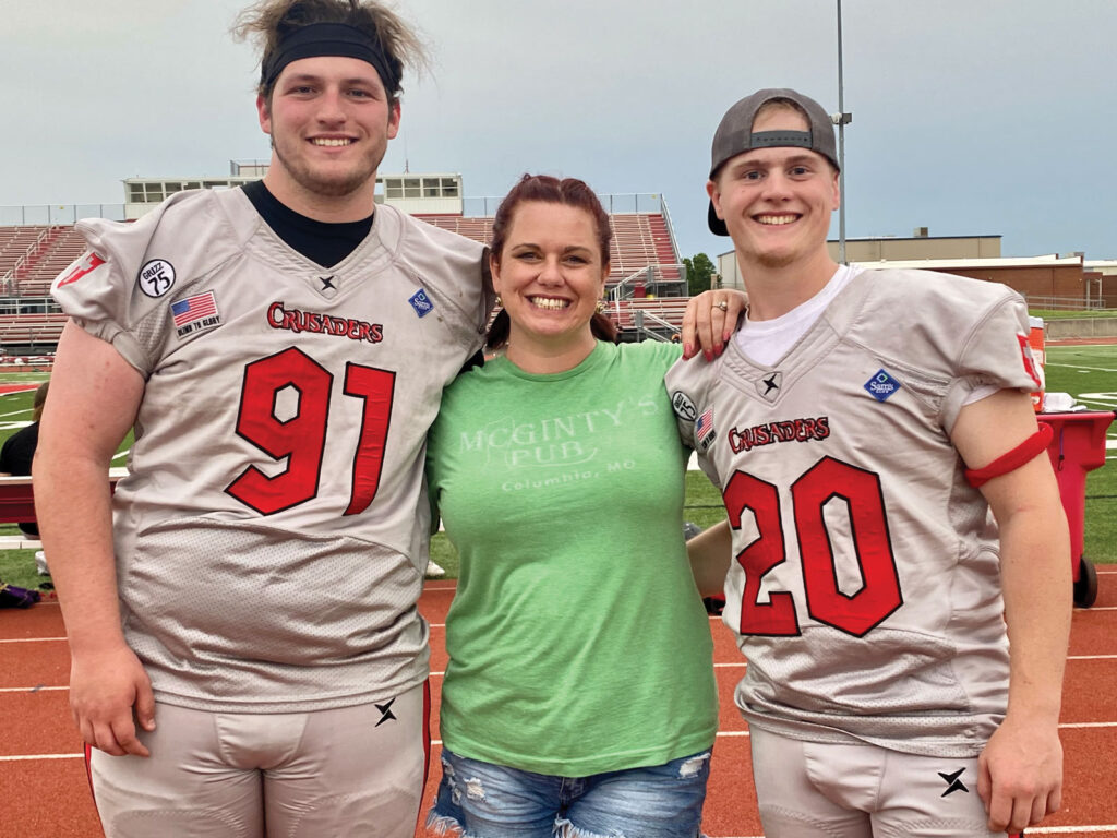 a mom stands next to her two sons in football uniforms by the side of the football field