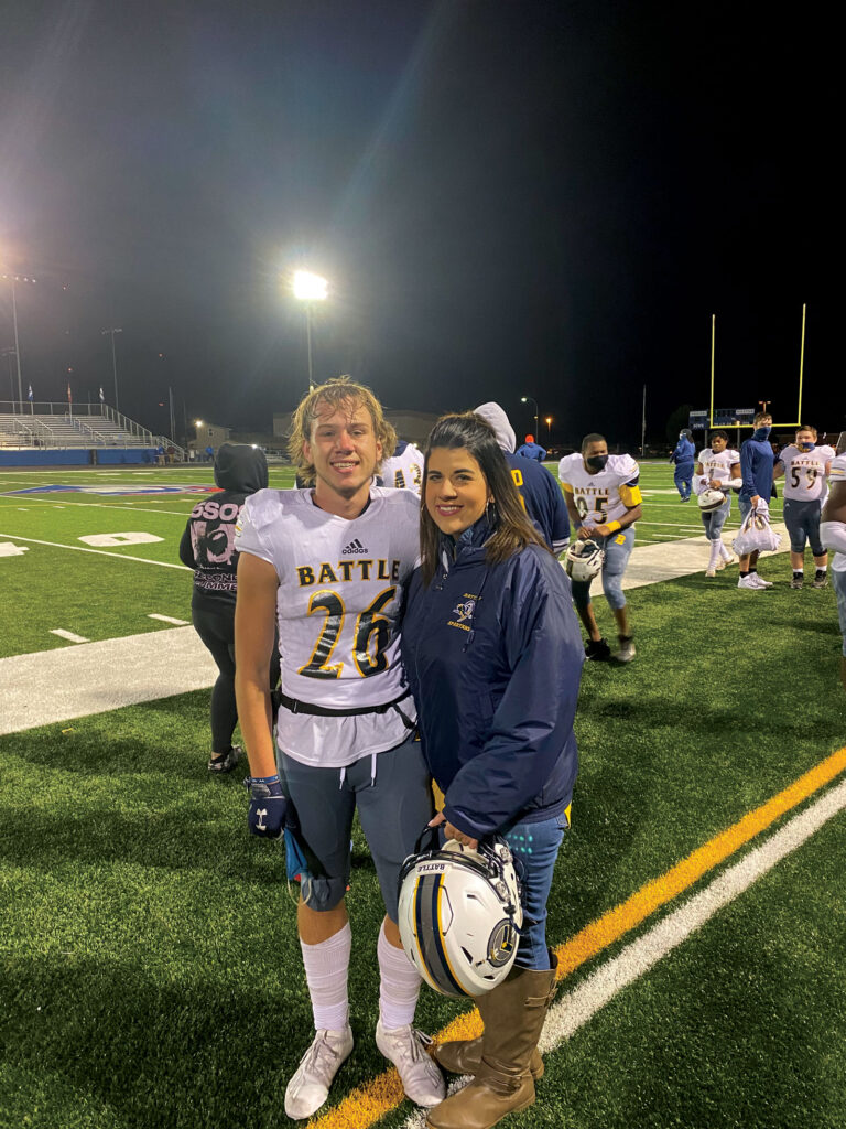 a women stands by her son in his football uniform by the side of the football field