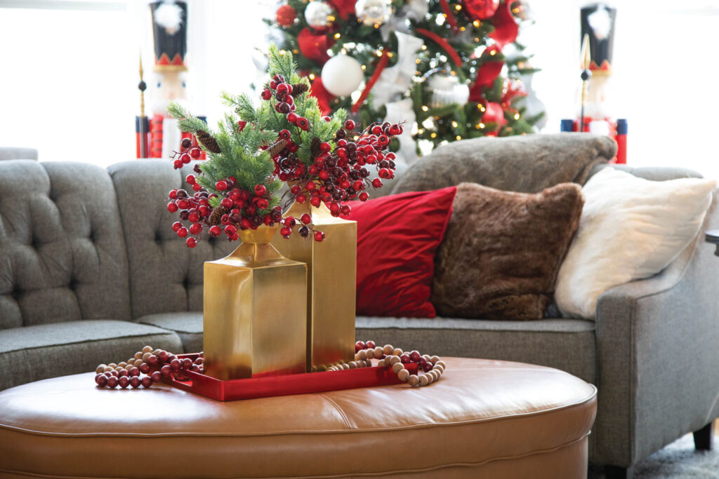 two gold vases with red berries and greenery in them sitting on a camel colored ottoman