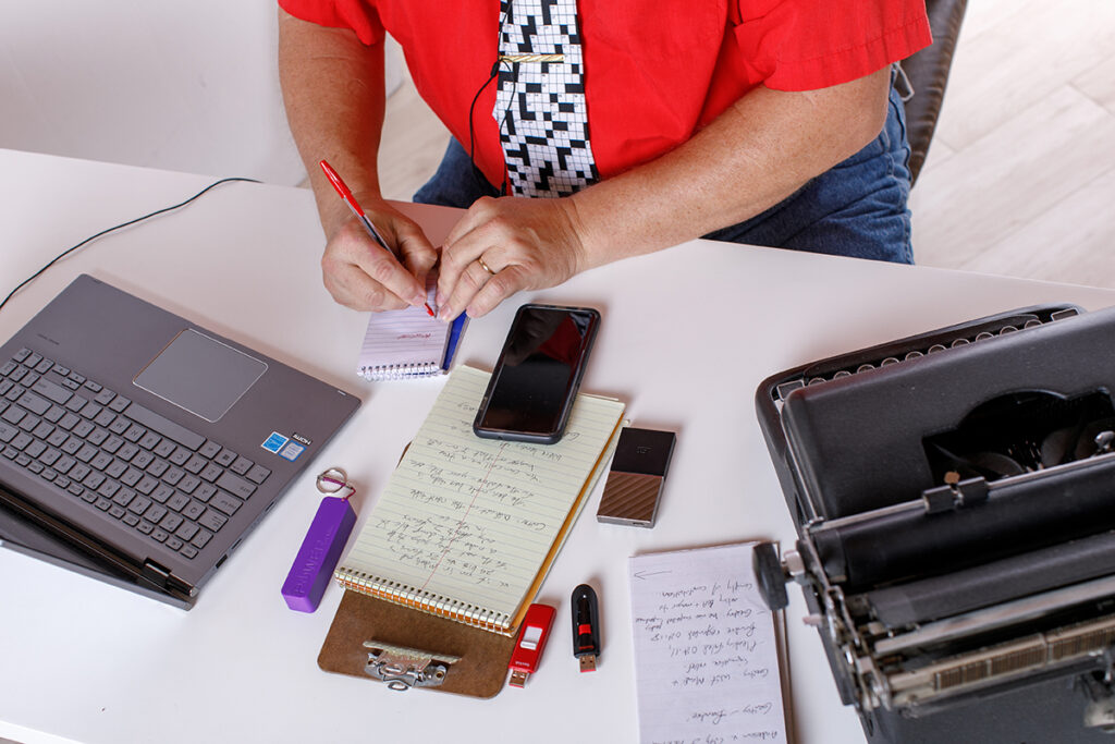 A man sits at a desk writing on a notepad surrounded by office supplies including a laptop, typewriter, notepad, phone, USB drive, and more