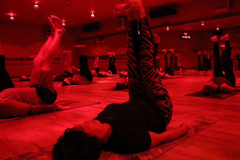 a group of people with legs raised participating in sauna yoga under red heat lamps