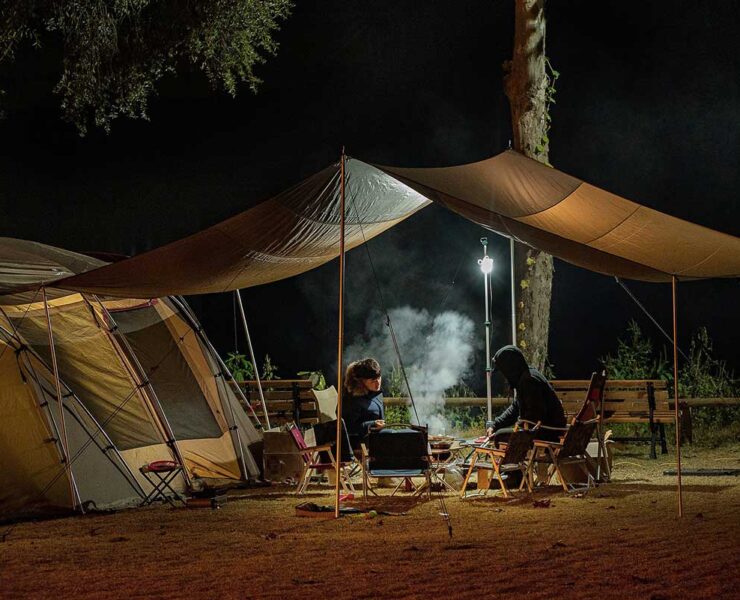Campsite photo showing two people relaxing under a canvass overhang between two tents, at night.