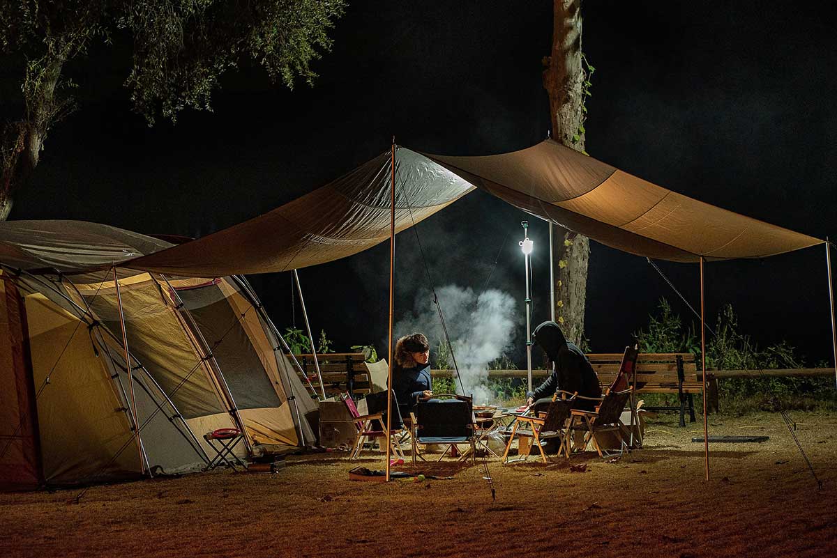 Campsite photo showing two people relaxing under a canvass overhang between two tents, at night.
