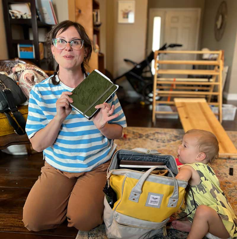 Jennifer Book Haselswerdt, wearing a blue and white striped short sleeve polo, smiles as she holds her Scribbles That Matter journal. Jennifer's toddler is searching her mom's Hmawari Backpack.
