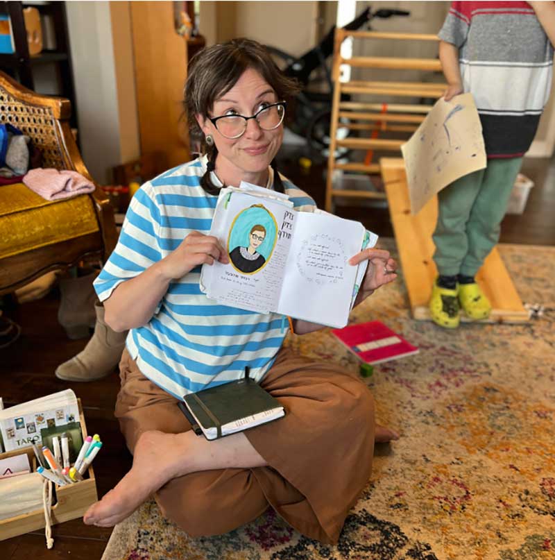 Jennifer Book Haselswerdt, wearing a blue and white striped short sleeve polo, smiles as she holds her Scribbles That Matter journal. 