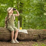A child dressed in khaki hiking clothes and a stiff brimmed hat looks through binoculars while seated on a large fallen log. He has a thermos and a map beside him on the log. There are green trees in the background.