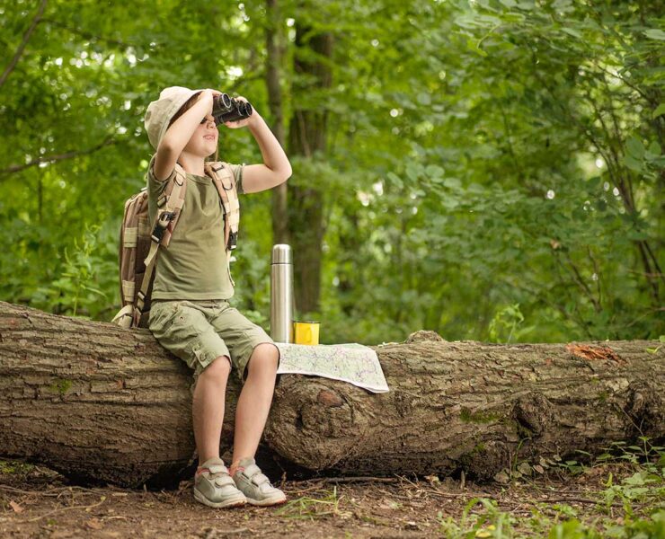 A child dressed in khaki hiking clothes and a stiff brimmed hat looks through binoculars while seated on a large fallen log. He has a thermos and a map beside him on the log. There are green trees in the background.