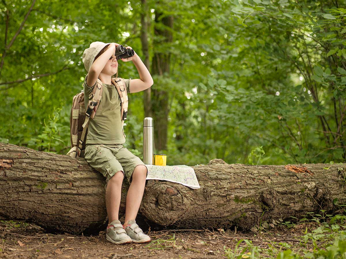 A child dressed in khaki hiking clothes and a stiff brimmed hat looks through binoculars while seated on a large fallen log. He has a thermos and a map beside him on the log. There are green trees in the background.