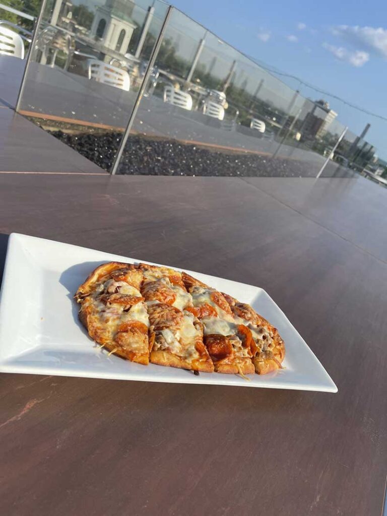 A flatbread pizza on a white plate with the downtown Columbia MO view in the background from The Roof.