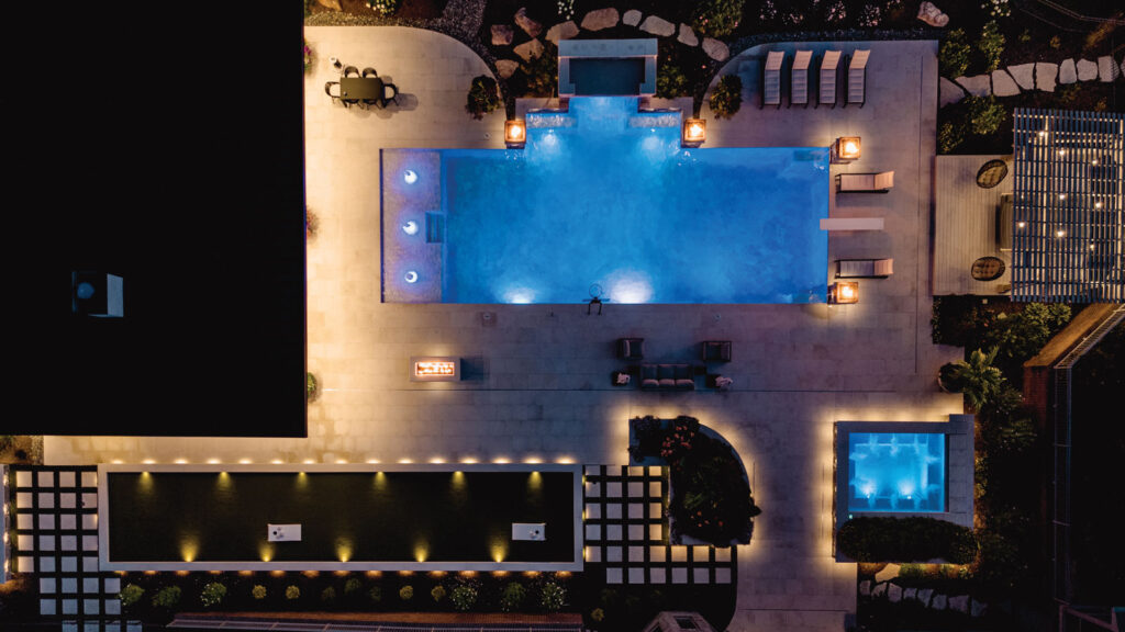 Aerial view of a backyard pool at night illuminated by accent lighting.