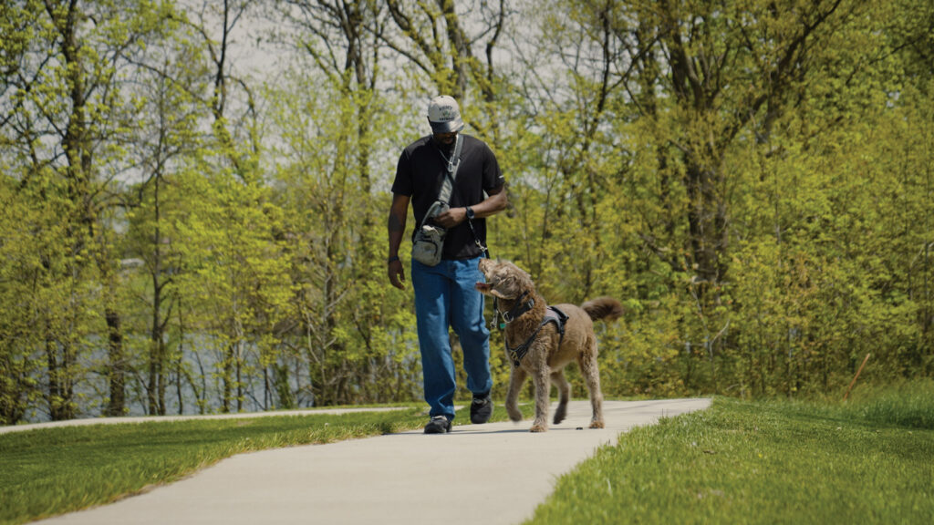 A military veteran and a trained service dog walk side by side along a paved trail.