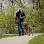 A military veteran and a trained service dog walk side by side along a paved trail.