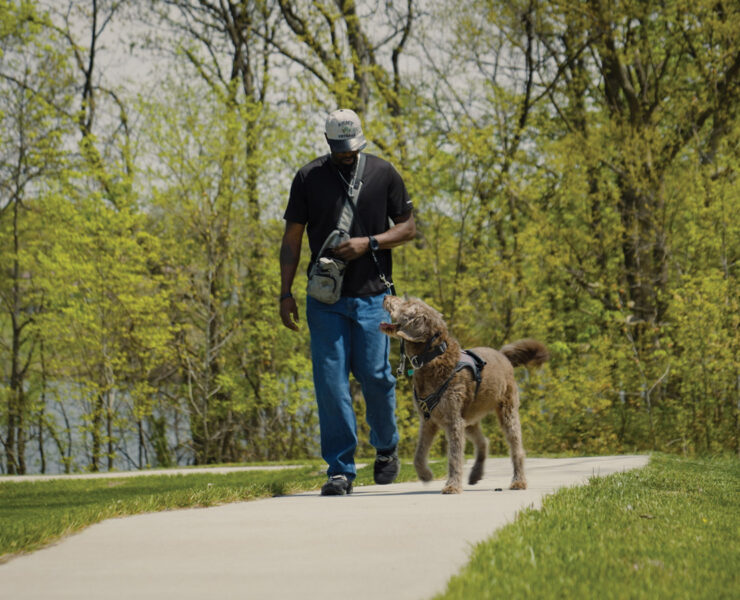 A military veteran and a trained service dog walk side by side along a paved trail.