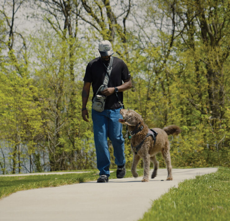 A military veteran and a trained service dog walk side by side along a paved trail.