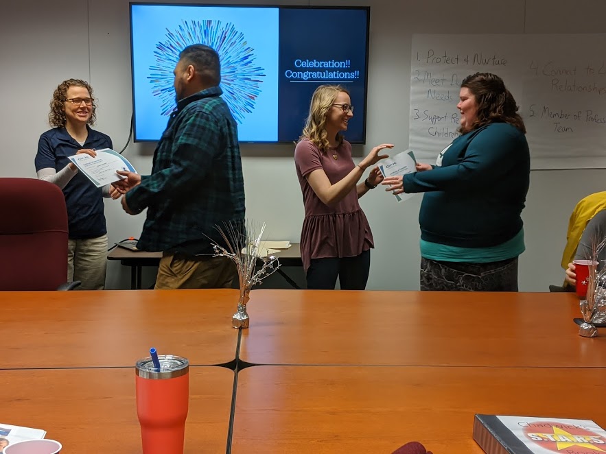 Four people are in a conference room exchanging certificates. A screen displays Celebration!! Congratulations!! on a colorful background. A table is in the foreground with drinks and papers. A flip chart with notes is on the right wall.