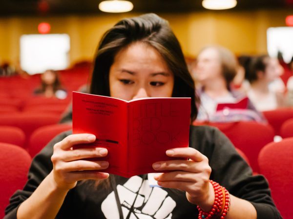 reading a red book titled Little Red Book. They have long dark hair and wear a black sweater with a fist graphic and red bracelets. The background shows blurred people and lights.