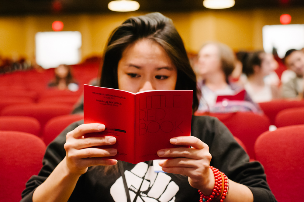 reading a red book titled Little Red Book. They have long dark hair and wear a black sweater with a fist graphic and red bracelets. The background shows blurred people and lights.