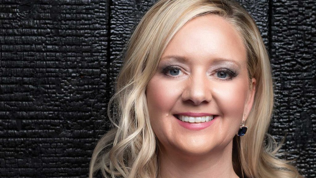 A woman with blonde hair smiles in front of a black textured background. She is wearing dark earrings and subtle makeup.