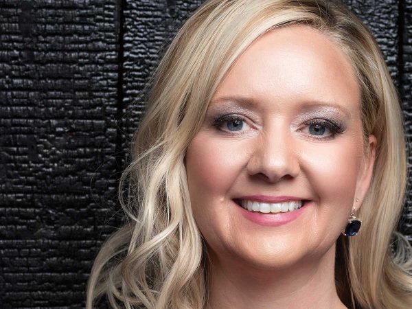 A woman with blonde hair smiles in front of a black textured background. She is wearing dark earrings and subtle makeup.