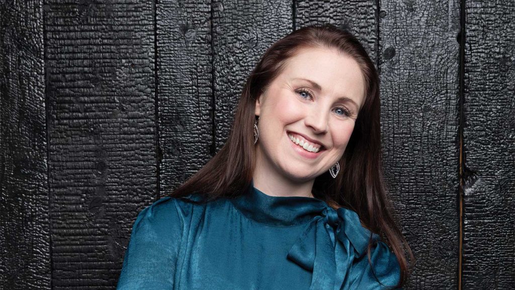 A woman with long brown hair smiles warmly at the camera. She is wearing a shiny teal blouse with a bow at the neck and silver earrings. The background is a textured black wooden wall.