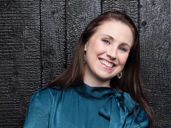 A woman with long brown hair smiles warmly at the camera. She is wearing a shiny teal blouse with a bow at the neck and silver earrings. The background is a textured black wooden wall.