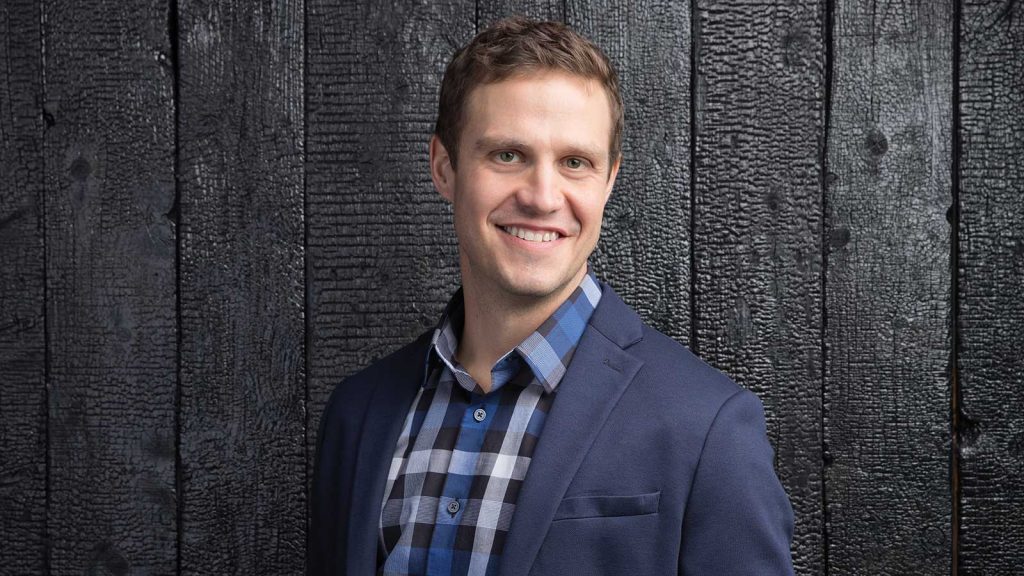 Smiling person wearing a blue blazer and checkered shirt stands in front of a textured dark wooden wall.
