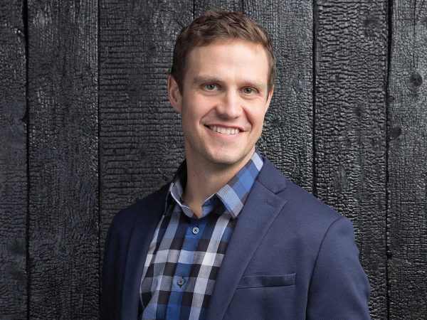 Smiling person wearing a blue blazer and checkered shirt stands in front of a textured dark wooden wall.