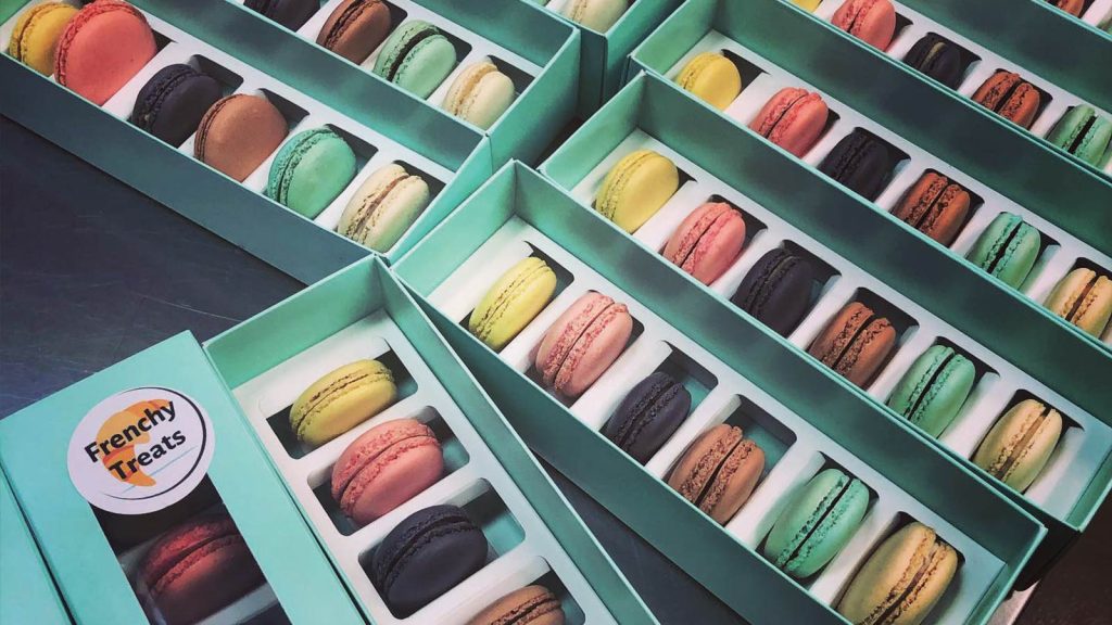 Boxes of colorful macarons in various flavors are neatly arranged in rows. Each box has a transparent lid and compartments for individual macarons. A label on the box reads Frenchy Treats.